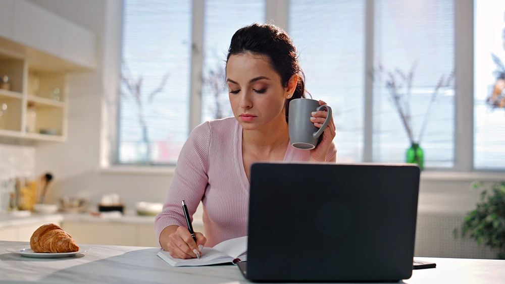 Woman doing self-paced online training in her kitchen, writing notes in front of a laptop.